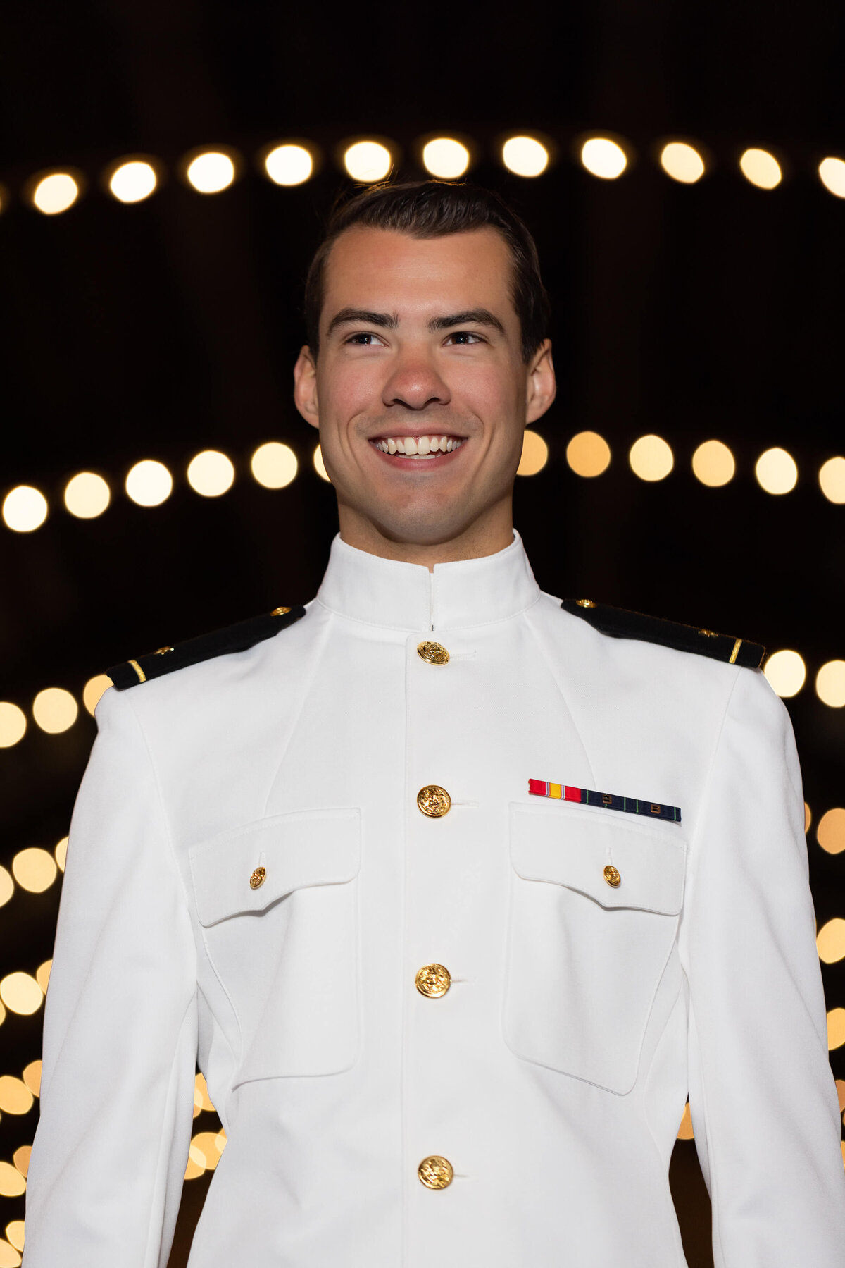 Naval Academy senior smiles in a portrait with the lights from Dahlgren Hall.