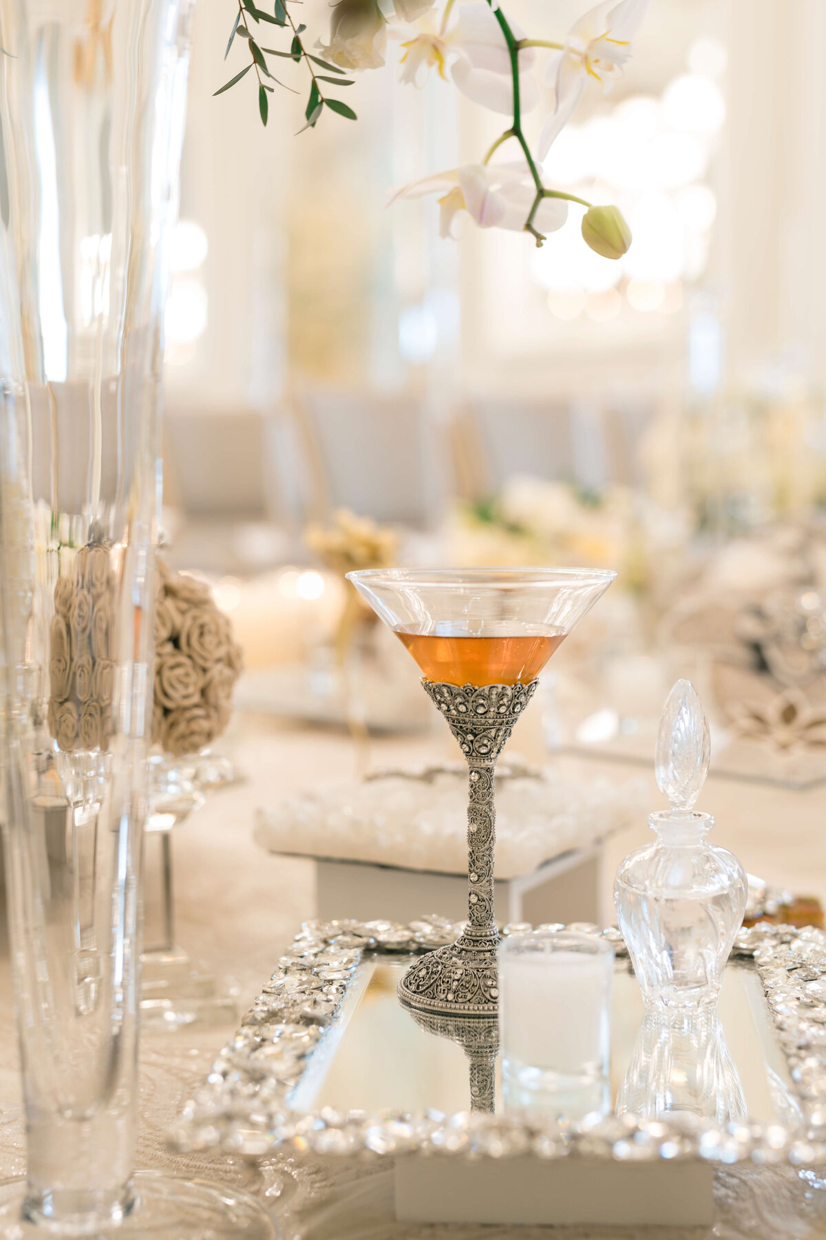 Elegant table setting with a decorative silver tray, featuring an ornate glass filled with a light orange drink. Surrounded by delicate white flowers and soft lighting, creating a sophisticated and luxurious atmosphere.