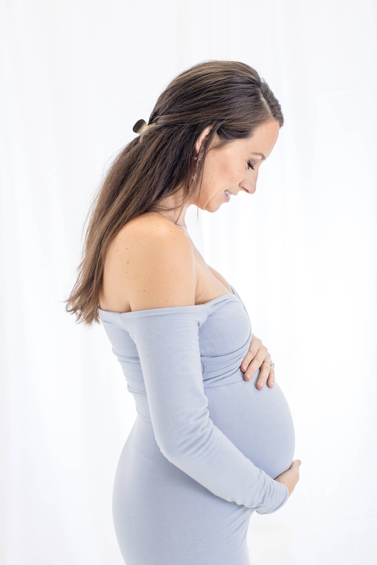 A pregnant person stands in profile against a white background, gently cradling their belly with both hands. They wear a form-fitting, off-the-shoulder, light blue dress, and their long, dark hair is tied back.