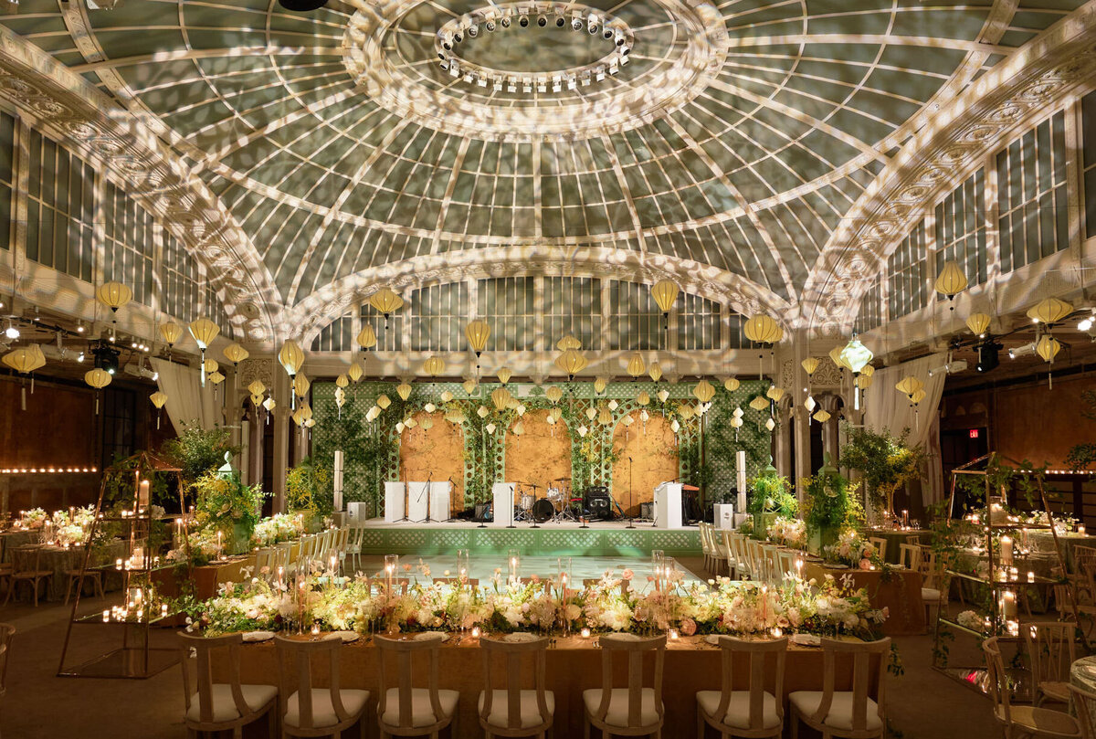 This stunning grand ballroom, set for a wedding, beautifully captures the essence of romance and luxury. Located at the iconic New York Public Library in New York City, this enchanting setting was expertly captured by photographer Shawn Connell and videographers Aaron Navak Films. The elegant tables adorned with candles, flowers, and greenery complement the ornate paneling and lush plants, creating a picture-perfect backdrop. The glass dome ceiling with intricate lighting further enhances the ambiance, casting warm reflections throughout the space.