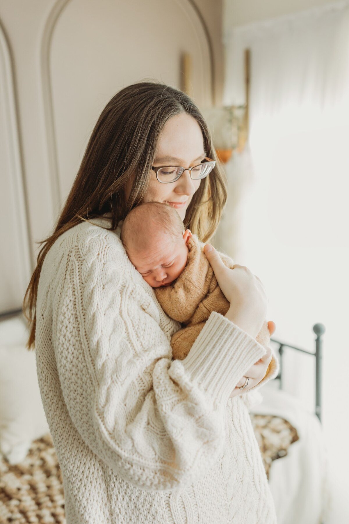 mother gently holds her sleeping newborn against her chest. She wears a chunky, cream-colored sweater and has long, brown hair. The soft, natural light from a nearby window creates a warm atmosphere in the room.