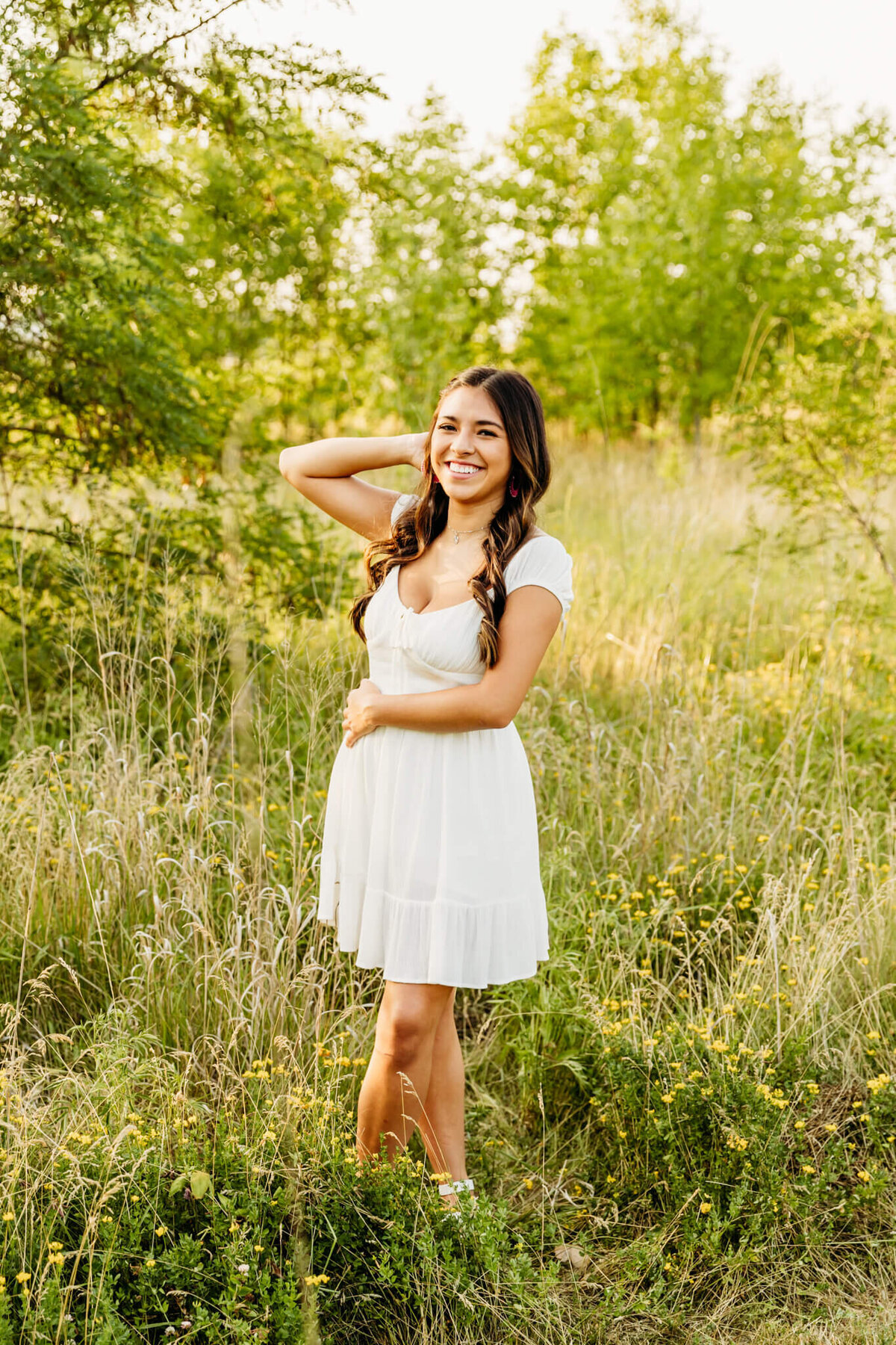 gorgeous teen girl in a white dress with ankles crossed smiling from ear to ear as she enjoys her senior photo session