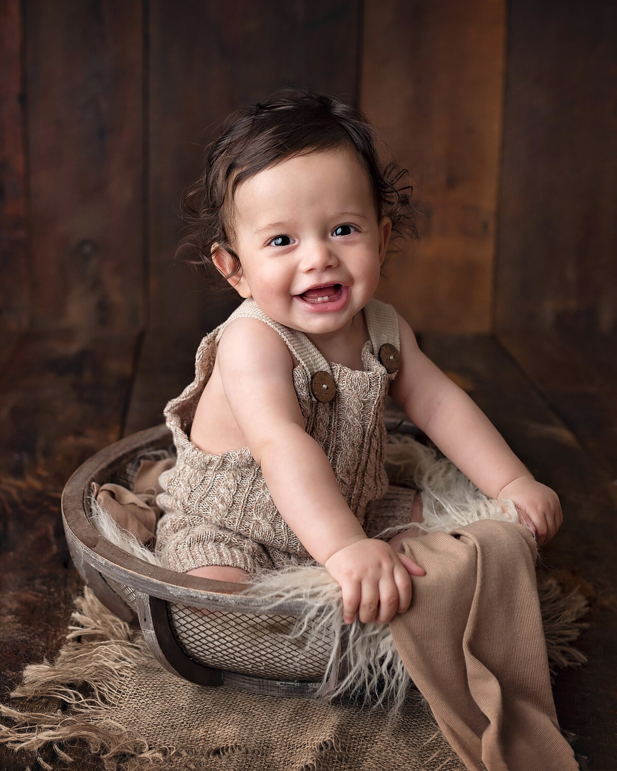 6-month milestone photoshoot at top West Palm Beach photography studio. Baby boy wearing a taupe knit jumper is sitting on furry blankets in a metal and wood basket. Baby is holding the side of the basket with a big toothy smile for the camera.