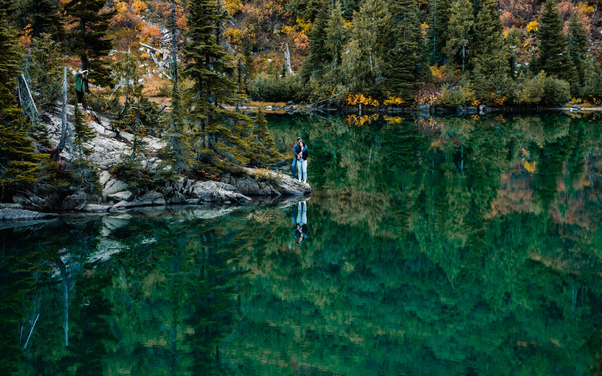 on the morining of their Washington elopement, a couple embraces on the rocky shore of a glacial lake surrounded by mountains, the fall colors are reflected in the lakes still watters