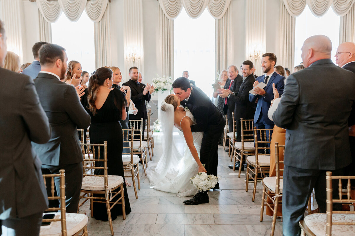 The newlyweds kiss in the center of the aisle as guests cheer around them, a joyful recessional moment documented by a Philadelphia wedding photographer.