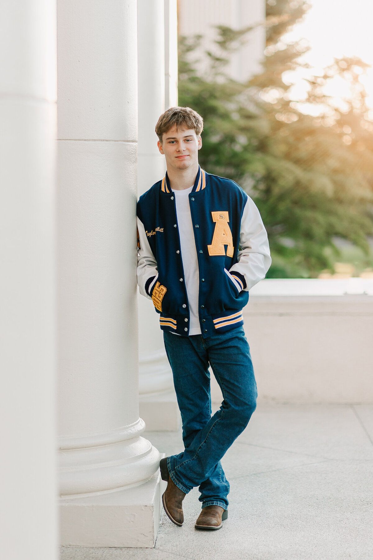 A senior boy stands leaning on a column downtown Albany, OR in a  varsity letter jacket and jeans