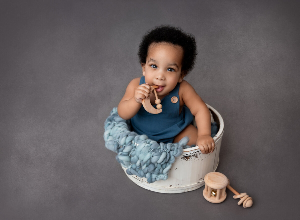 Baby boy sitting in white bucket playing with wooden toys for 6-month baby photo session.