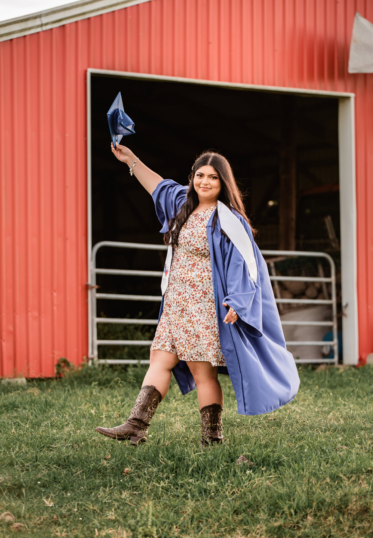 A senior holds her blue cap in the air in front of a red barn in celebration of her upcoming graduation.