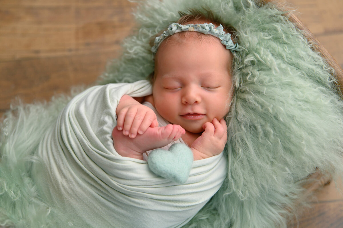 newborn girl wrapped in green holding a tiny green heart in a tiny bed for her newborn photos in Burlington