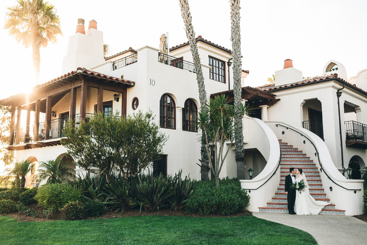 A photograph in color of a couple on their wedding day at The Ritz-Carlton Bacara in Santa Barbara, California. The photograph shows the bride and groom at the bottom of a curving staircase of a Spanish style villa. There is green grass and palm trees with the sun peeking out over the roof. Wedding photography by Stacie McChesney/Vitae Weddings.