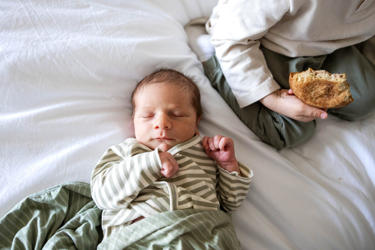 Baby boy sleeping on bed while his big brother kneels nearby eating a cooking
