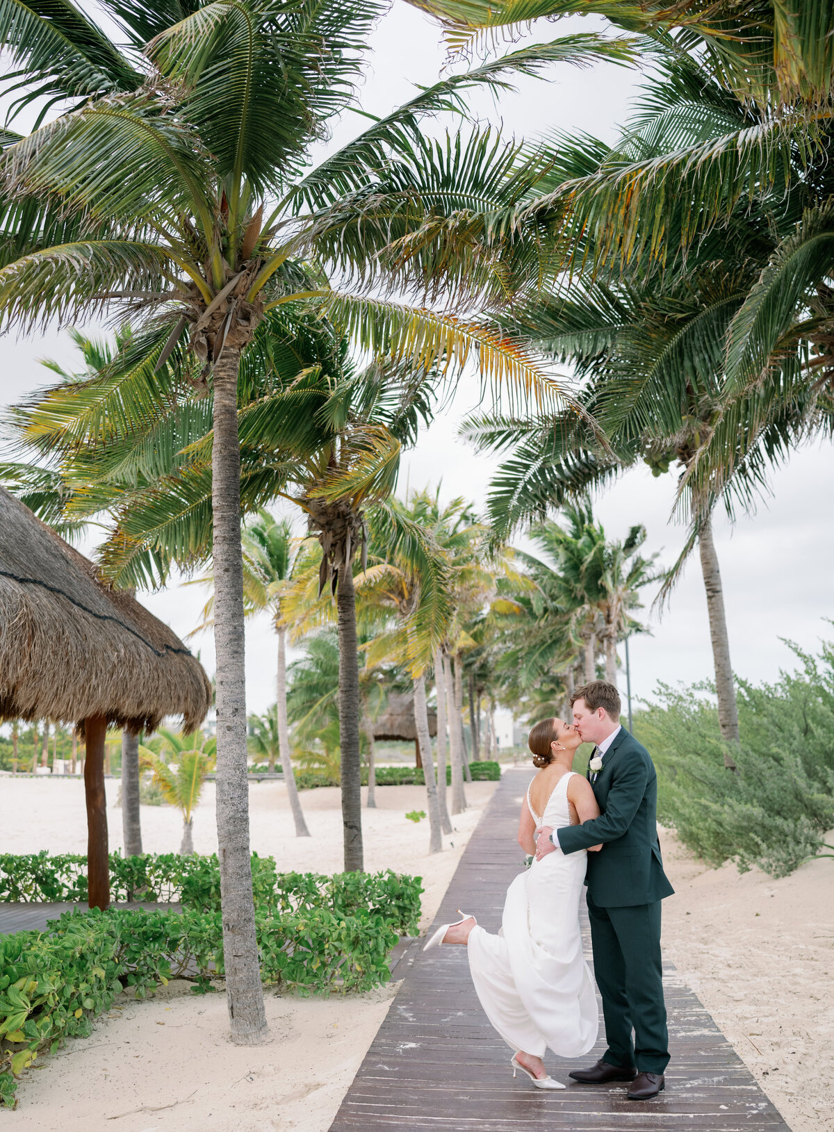 A groom kisses his bride while walking on a palm tree lined path near the ocean in Cancun