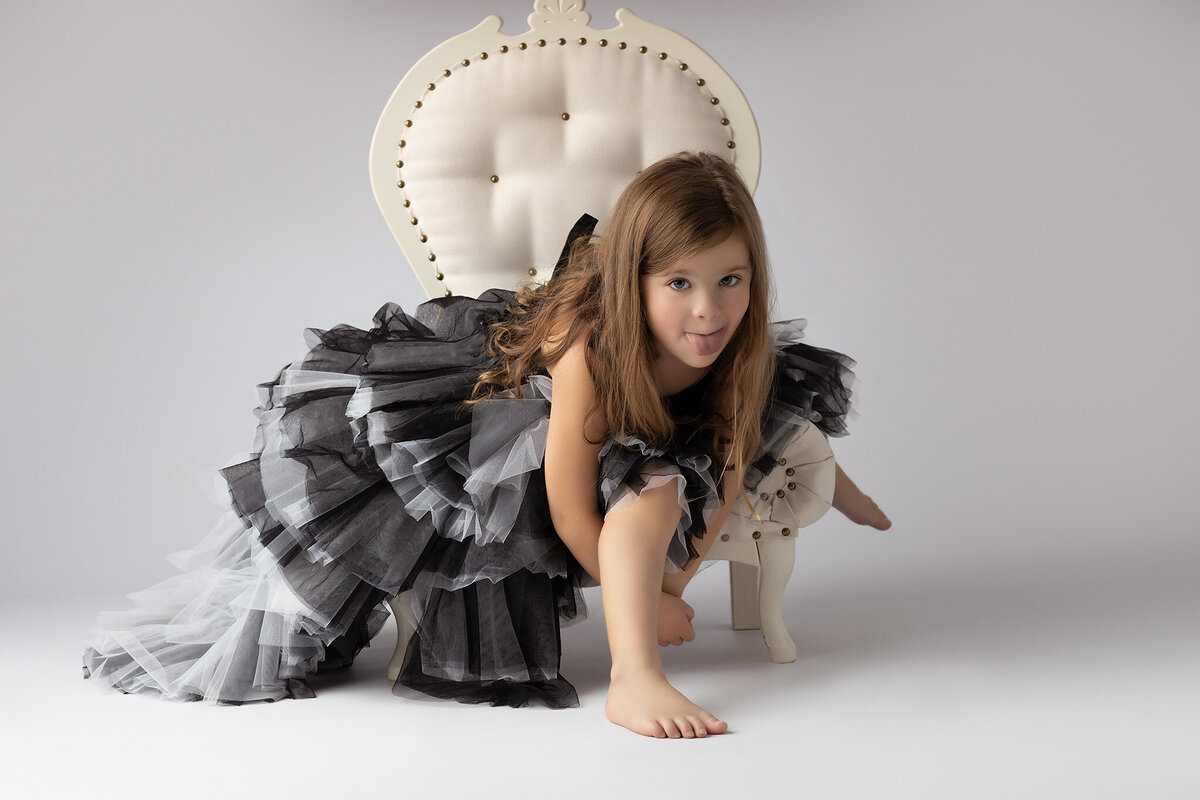 A playful girl in a black and gray ruffled gown sticking out her tongue while sitting on a stylish chair.
