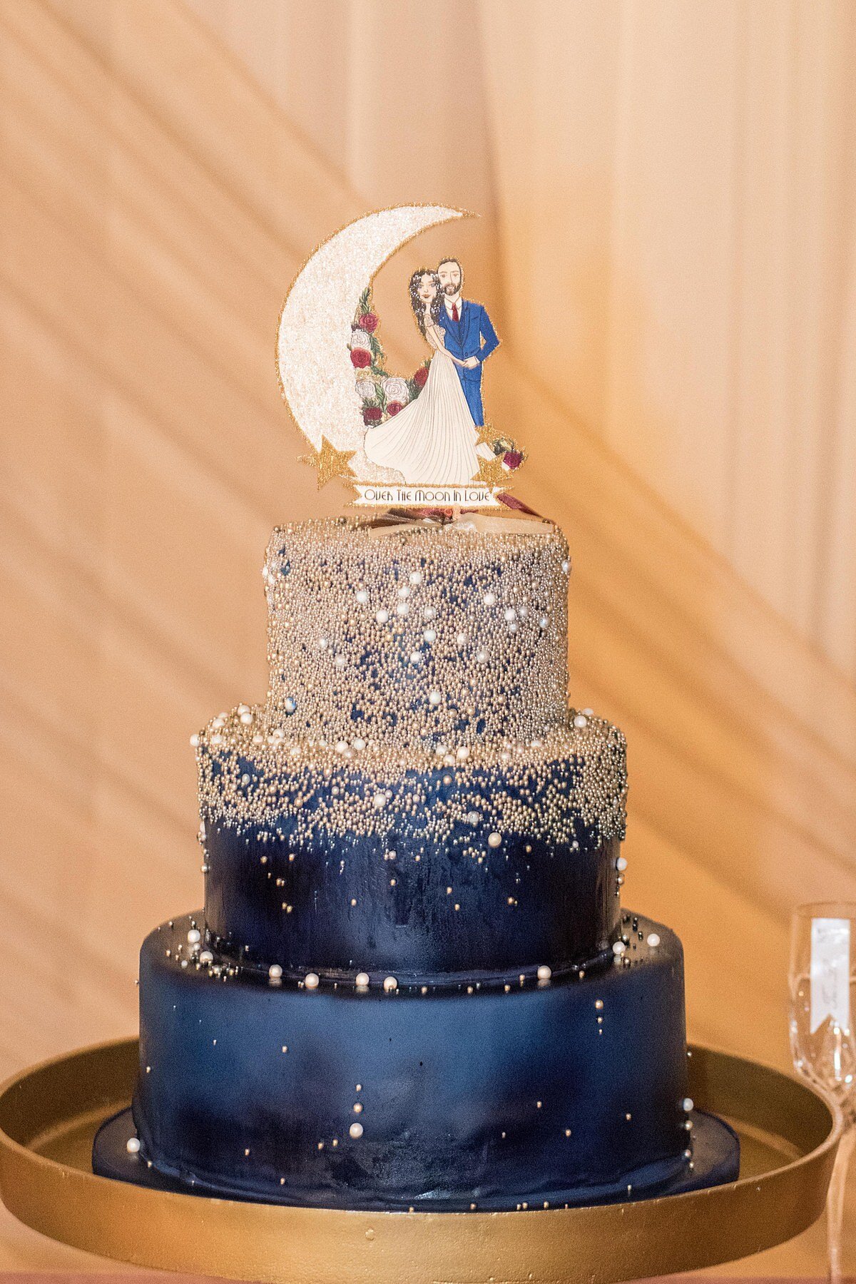 Navy blue  and gold three tiered 1920s inspired celestial wedding cake with bride and groom standing next to a crescent moon cake topper against a champagne gold drapery backdrop