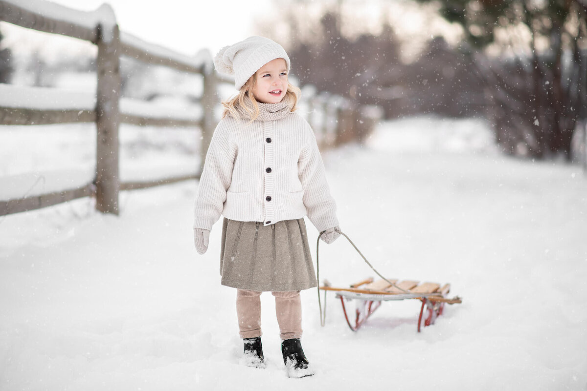 Young girl dressed in warm white layers in the winter in kamloops pulling a wooden sled.  Portrait by Brittany Danielle