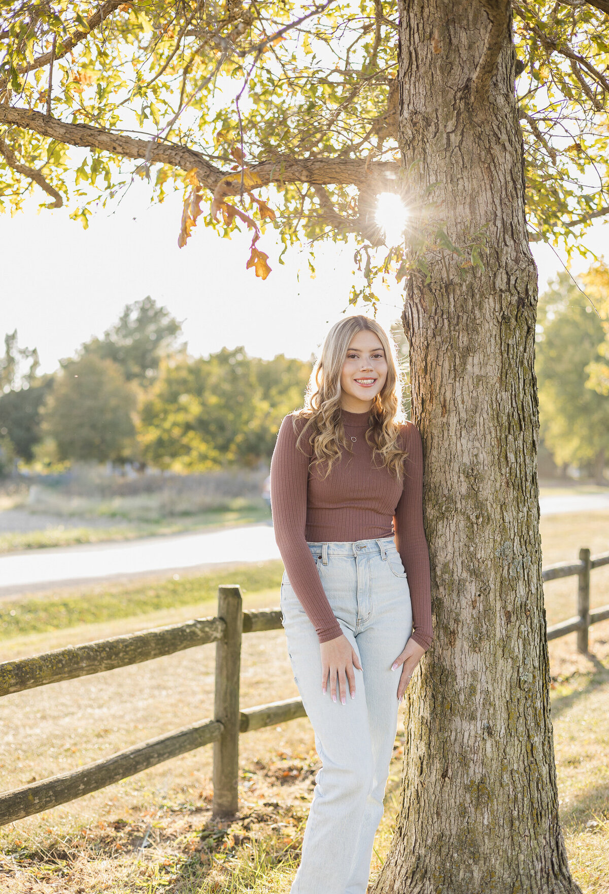 High school senior female with long blonde hair, leaning against a tree with the sun and trees behind her, wearing a long sleeve purple shirt, blue jeans, and smiling at the camera