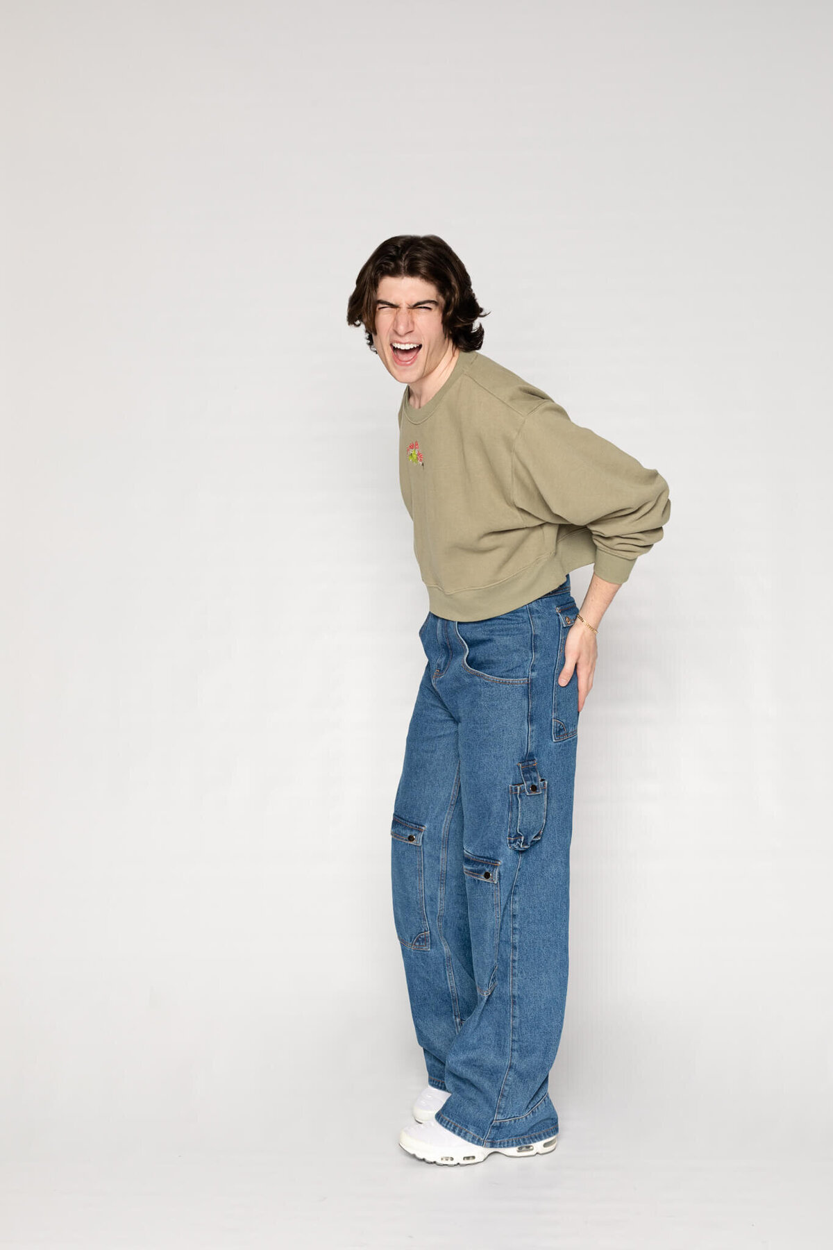 A person with shoulder-length hair is wearing a beige sweatshirt and wide-leg blue jeans. They stand against a plain background, laughing and standing in a playful pose with their right hand on their hip.