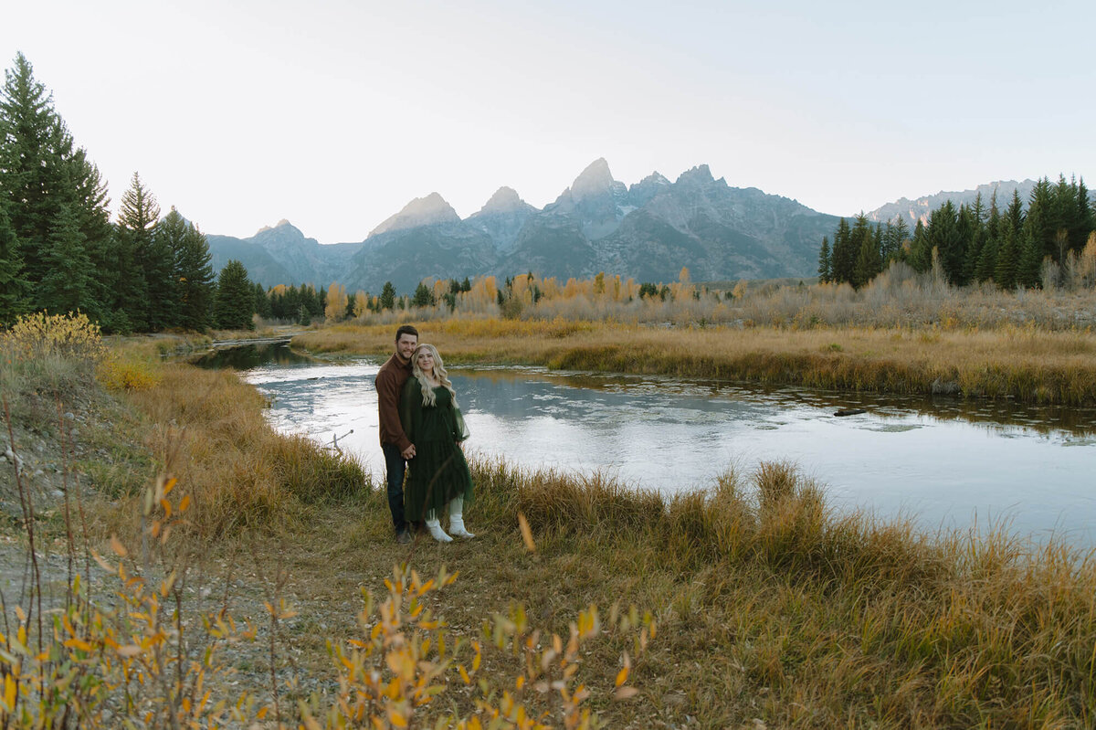 A woman leans back against her fiancé for an engagement shoot in Grand Teton National Park. Behind them is a river and in the far distance, the Tetons.