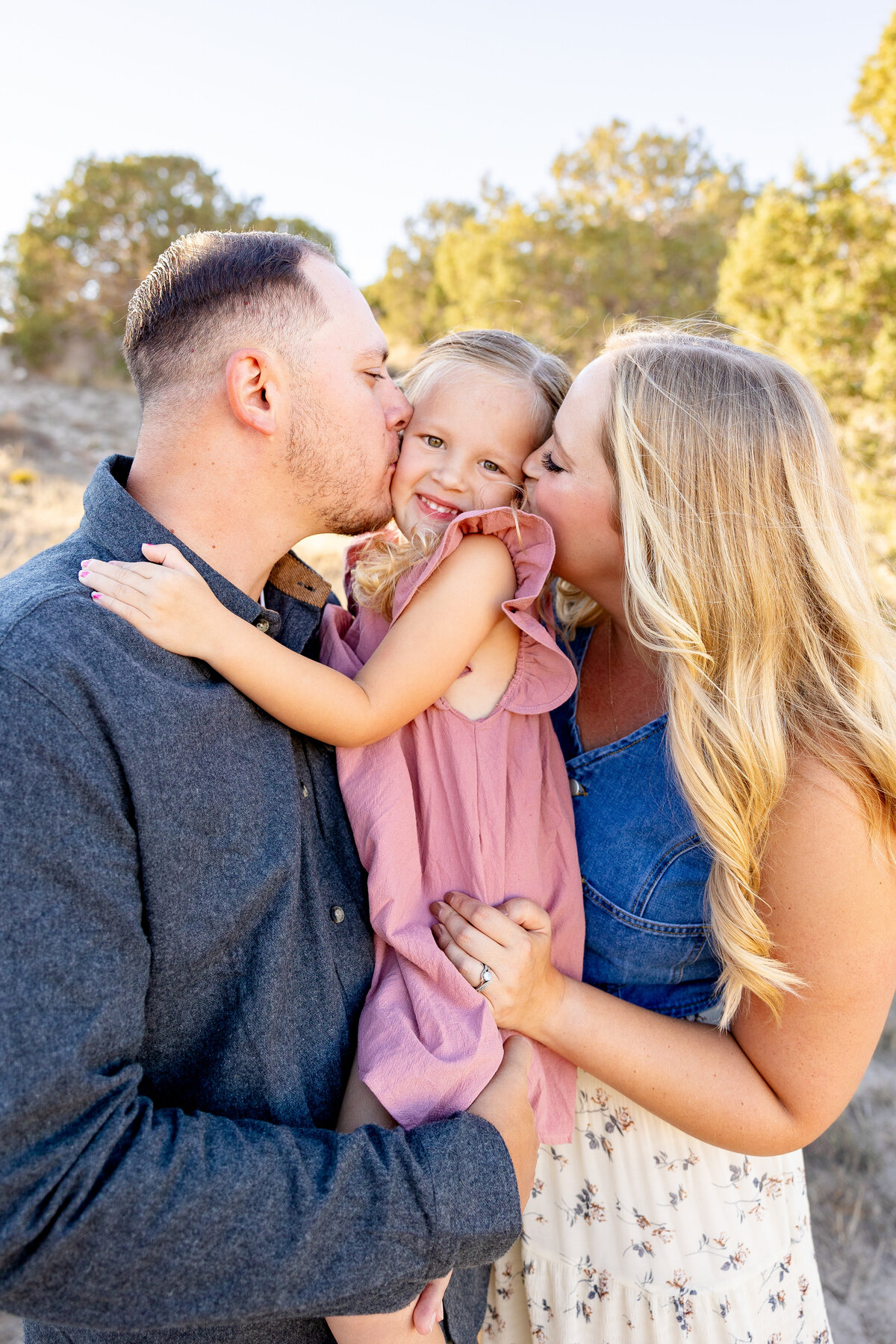 A little girl giggles as her parents kiss her cheeks, embracing her tightly in a loving moment. The background features rolling desert hills and golden fields.