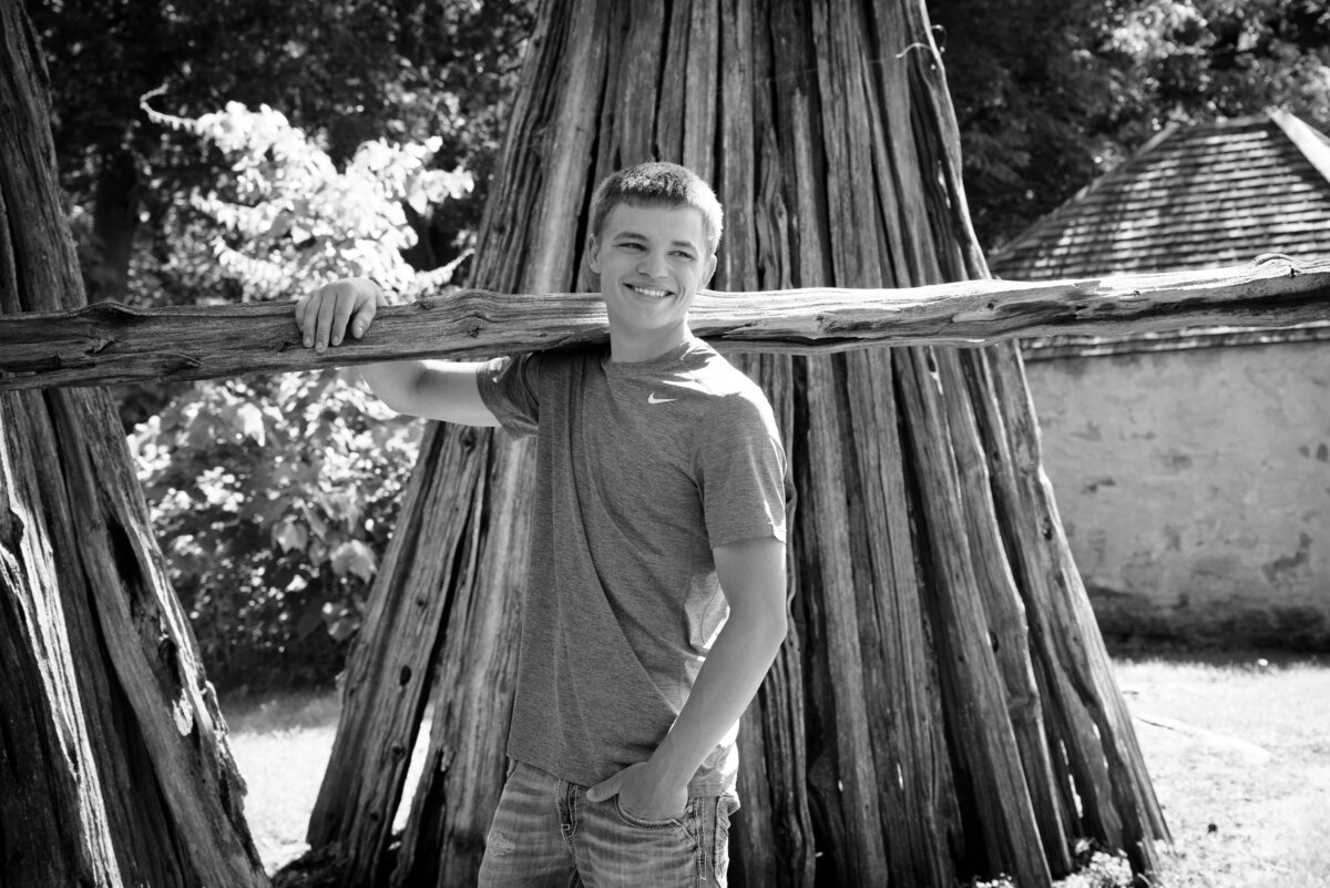 Black and white picture of Luxemburg Casco high school senior boy wearing a short sleeve blue t-shirt and jeans holding wooden fence post by cabin at Devil's River Campground near Green Bay, Wisconsin.