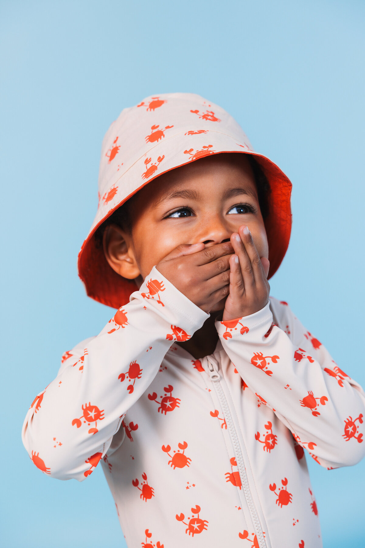 Smiling child wearing a Swim Essentials UV swimsuit and matching bucket hat with a red crab print, playfully covering their mouth while standing against a light blue background.