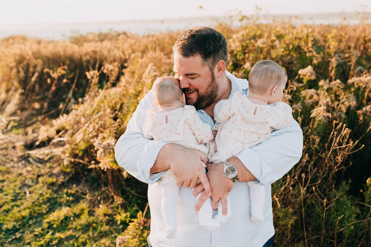 A father holds his twin baby girls in a field at sunset