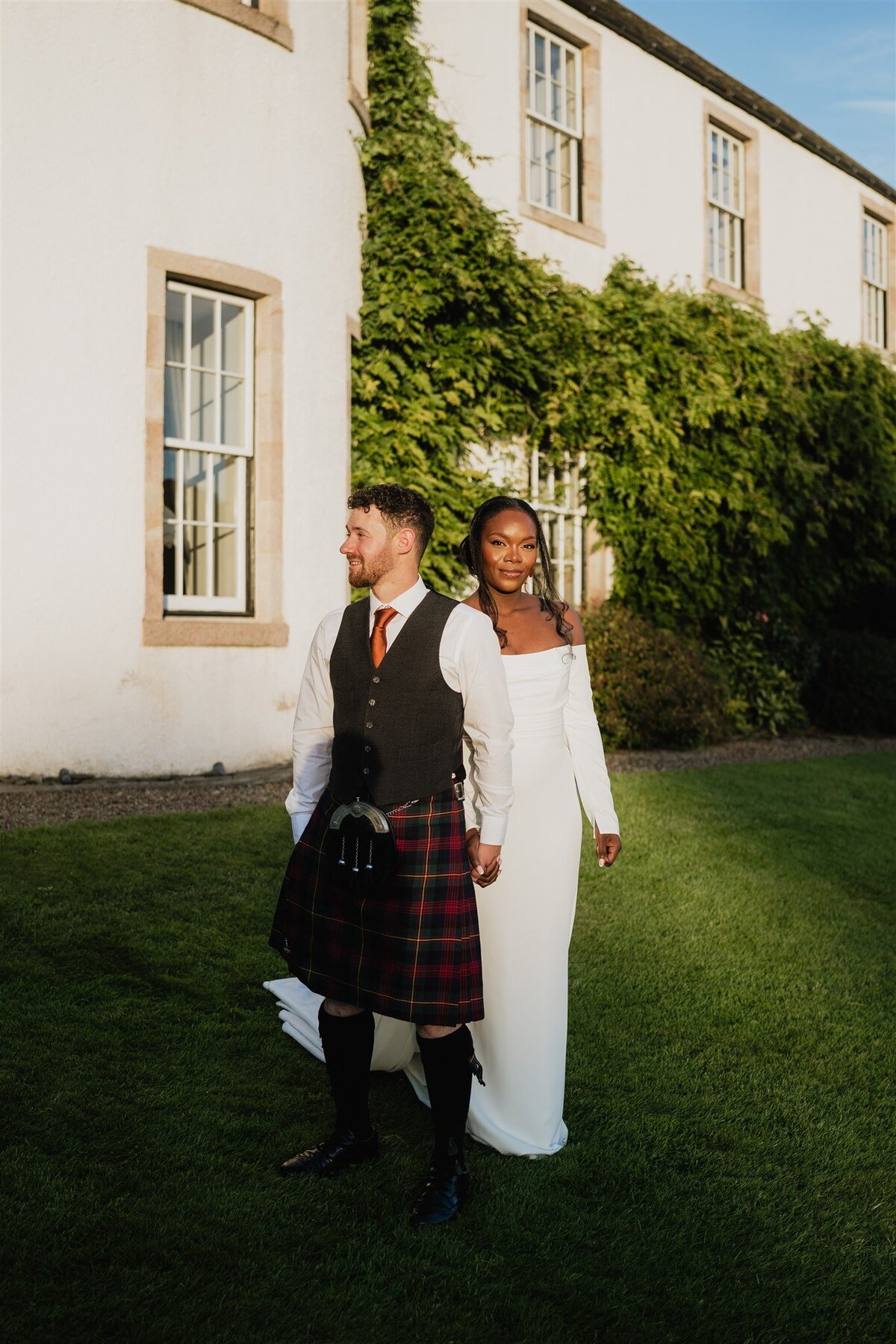 A bride and groom hold hands and pose for their wedding photos during golden hour. Logie Country House is seen being them. The bride wears a long white wedding dress and the groom wears a kilt. They look relaxed and look opposite directions.