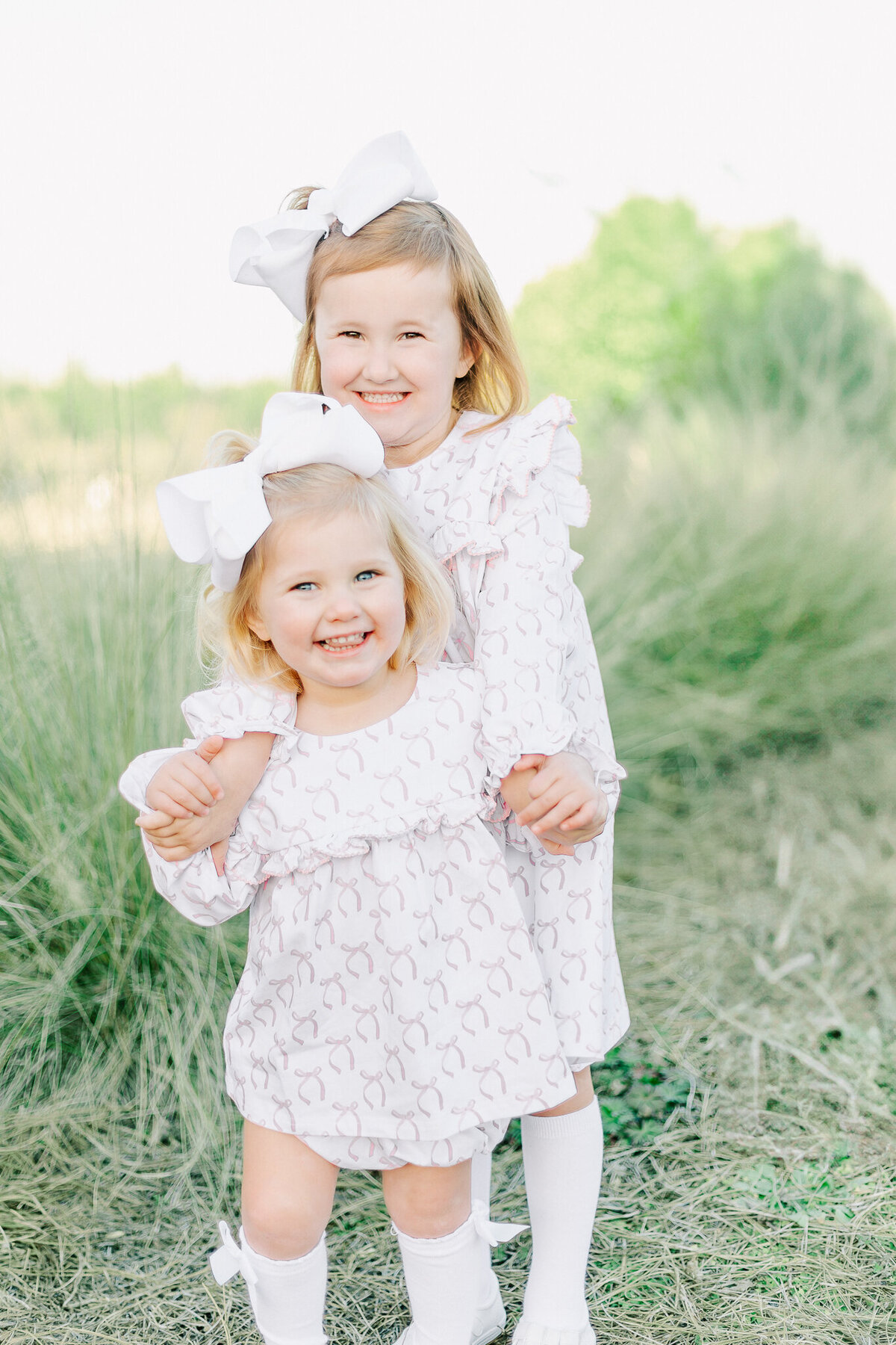 Two young sisters with matching white bows and pastel dresses smile brightly while embracing in a natural outdoor setting. Their joyful expressions and sweet bond capture the warmth and innocence of childhood, reflecting the light and timeless style of a Birmingham family photographer.