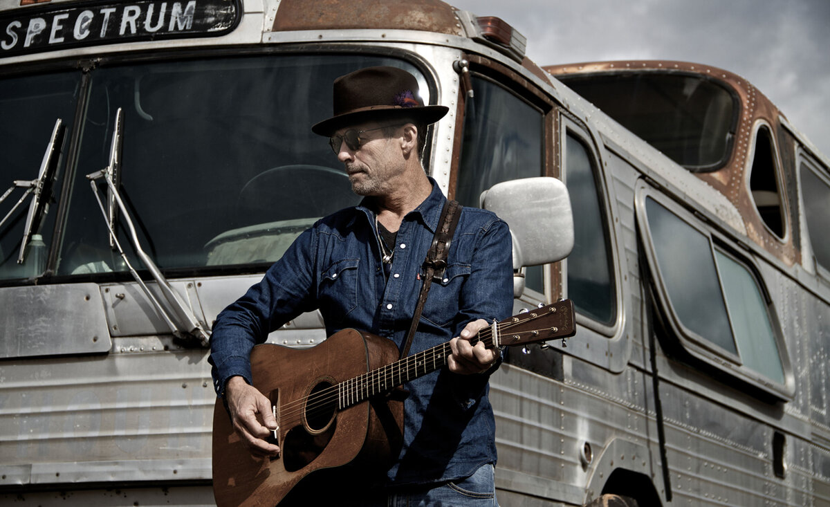 Male musician portrait Barney Bentall wearing blue denim shirt brown cowboy hat standing playing guitar old tour bus behind
