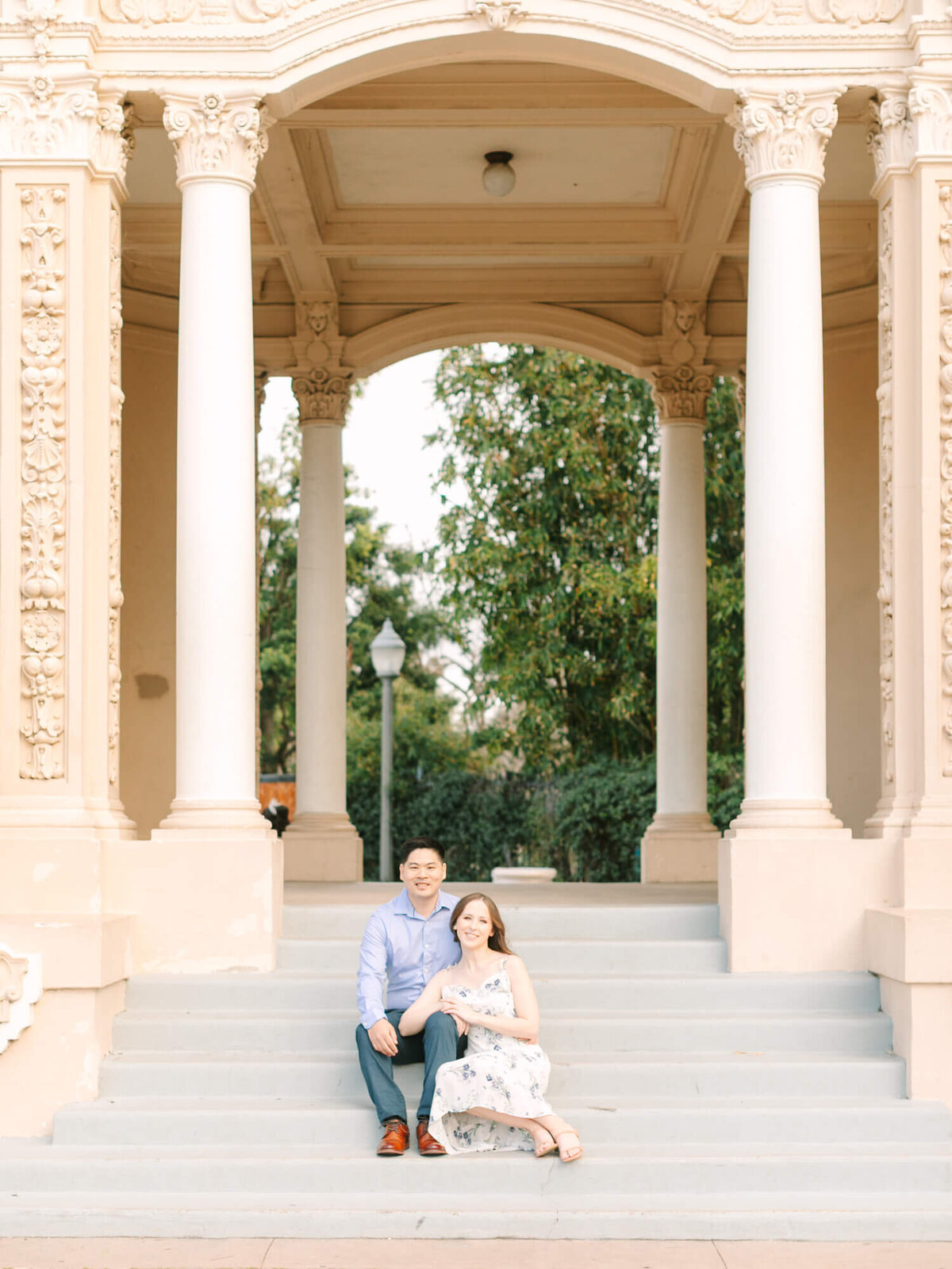 engaged couple sits together on wide stone steps under an ornate, columned archway with trees and a lamp post in the background