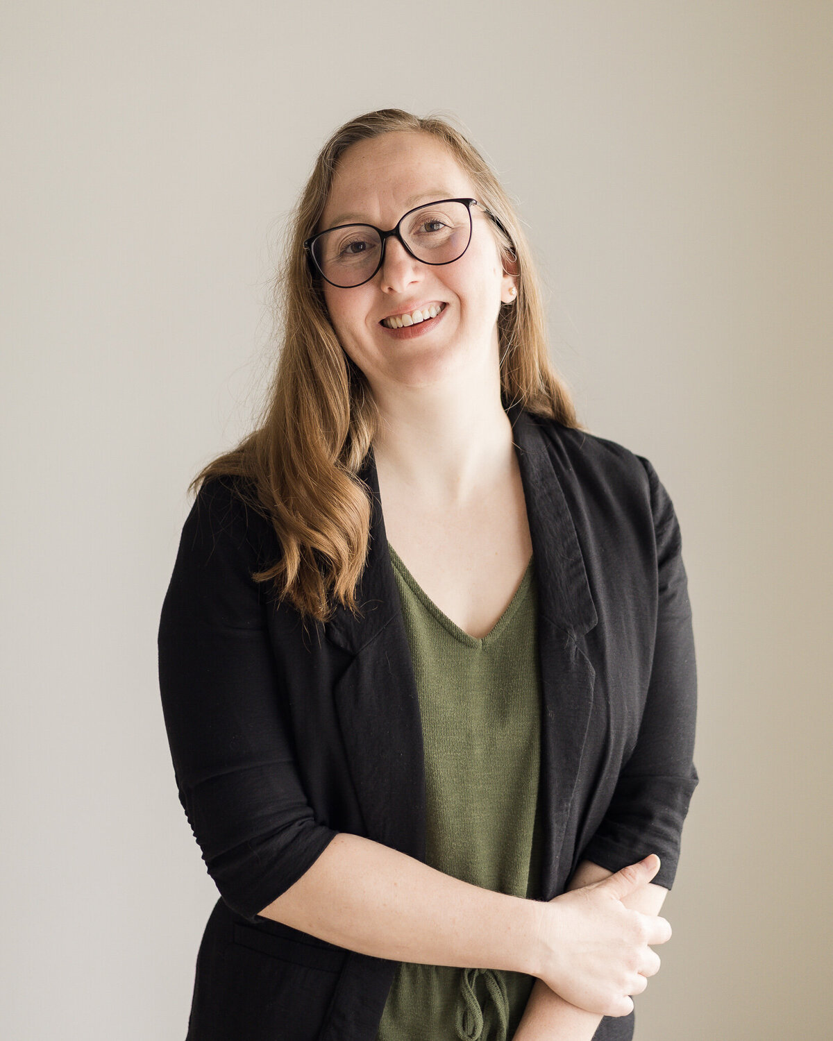 Strawberry blonde female, standing with one hand over her other arm, wearing a dark green shirt, with a black jacket over the green outfit, wearing glasses, smiling at the camera
