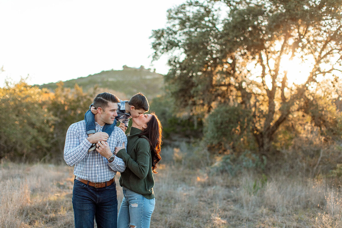 Child on father's shoulders kissing mother outdoors in golden light - Sara James San Antonio family photographer captures sweet three-person family moments and playful interactions in beautiful Texas natural settings