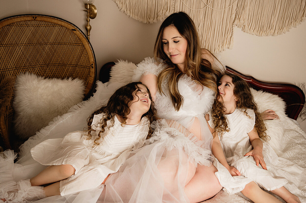 Mom expecting her third child cuddling with her twin daughters on a bed with boho macrame setup, by Tamara Danielle, Maternity Photographer in Greater Toronto area.