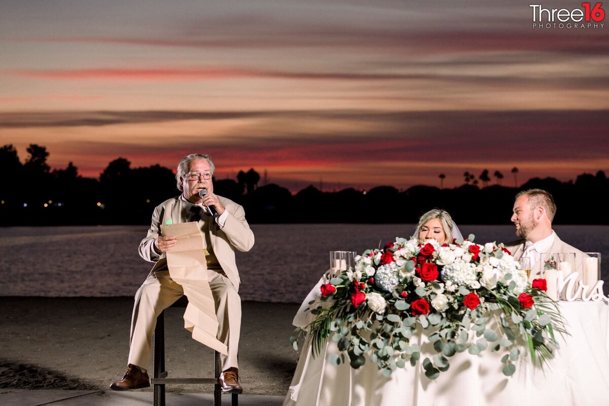 Bride and Groom laugh while sitting at the sweetheart table while her father toasts them