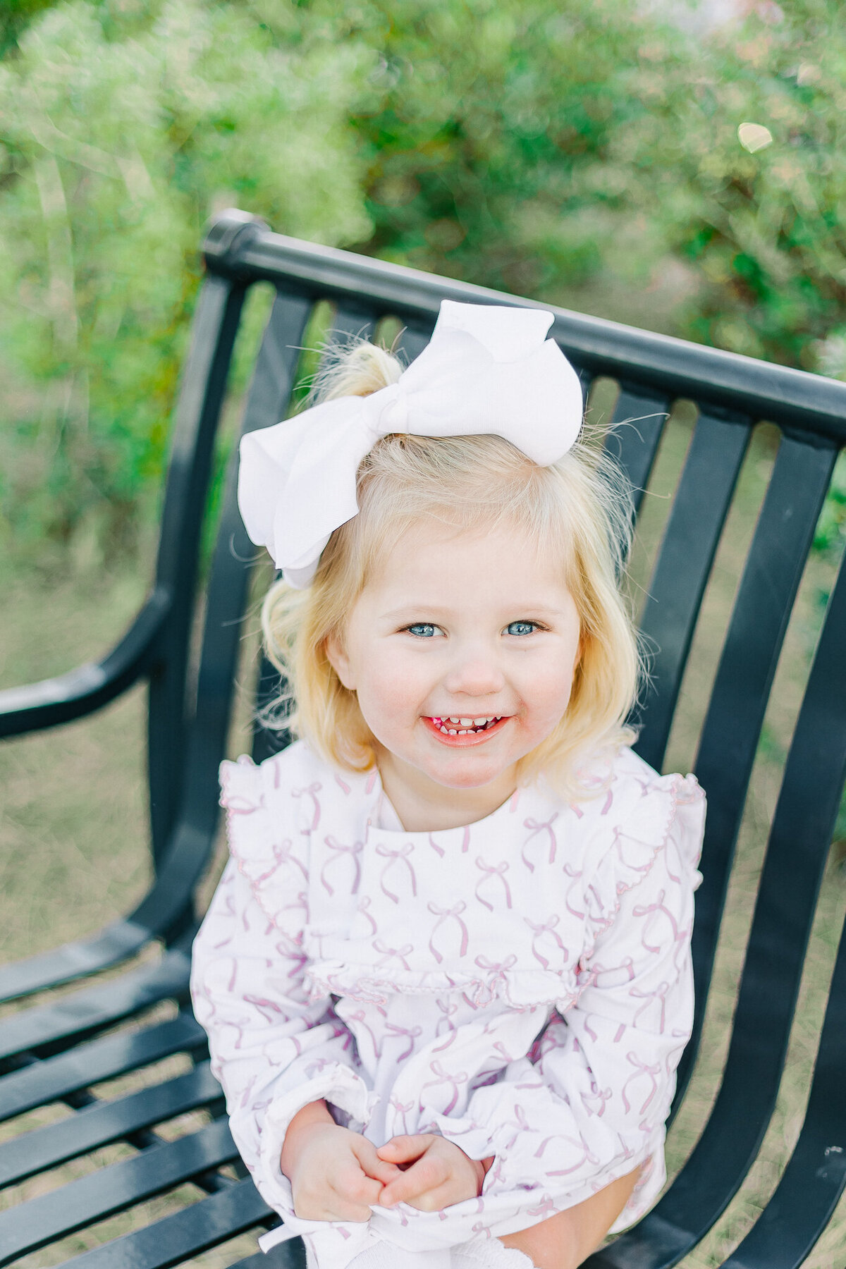 A young girl with blonde curls and a big white bow sits on a park bench, smiling brightly. Dressed in a classic ruffled dress, she radiates pure childhood joy, capturing the light and timeless style of a Birmingham family photographer.