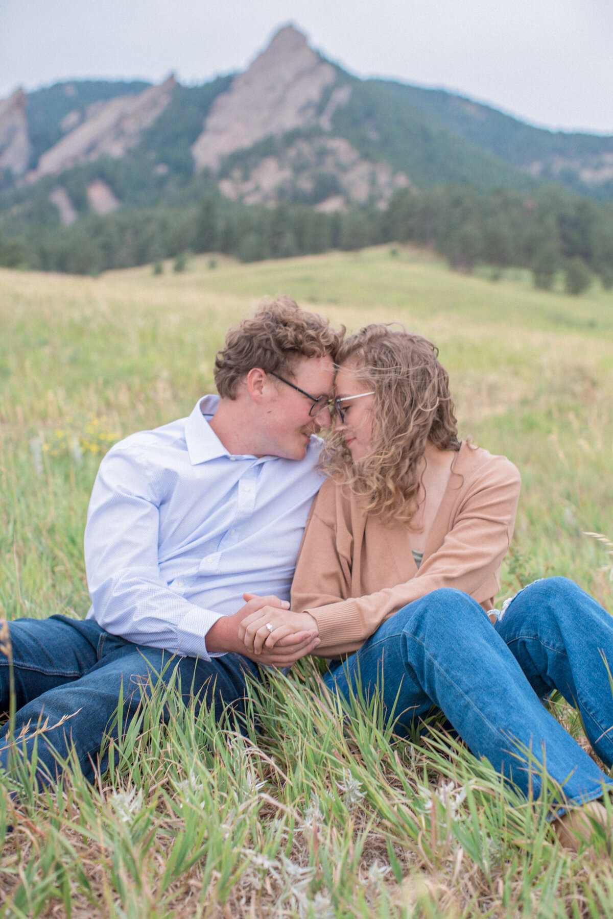 A newly engaged couple snuggle together in the a field with the Boulder flatirons in the background