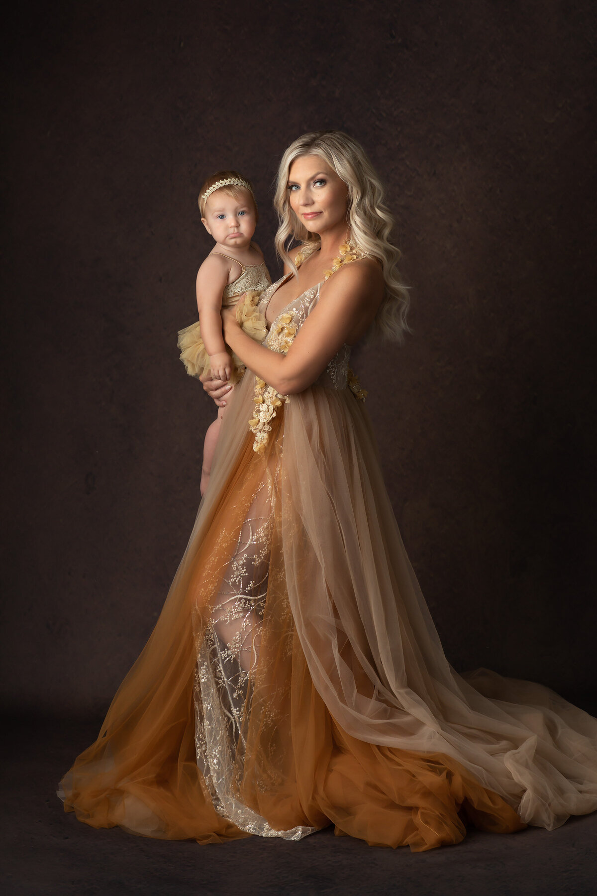 A mother and her baby share a loving embrace, both dressed in golden gowns, captured by a Dallas family photographer.