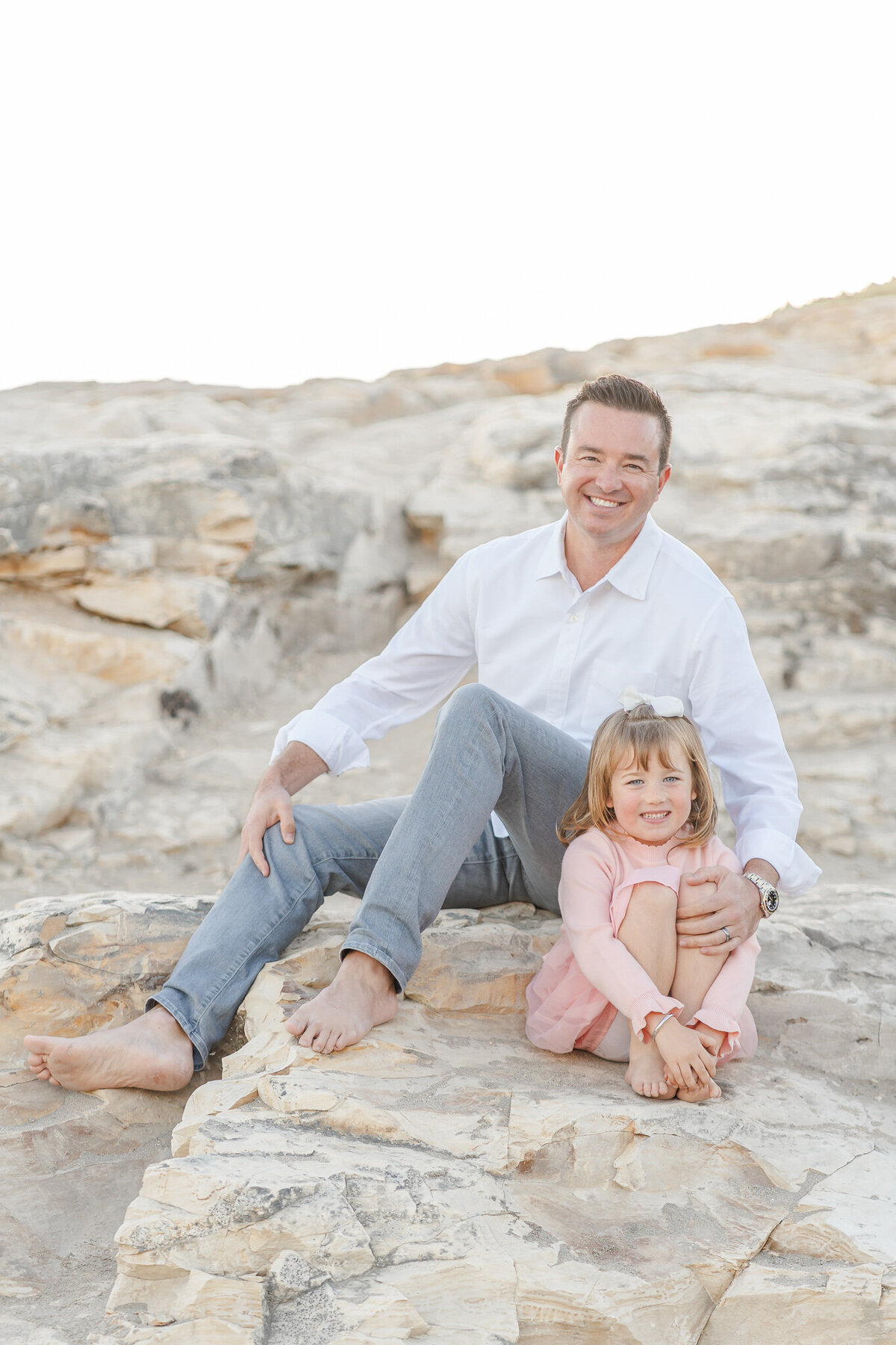 dad and daughter sitting together on a rock at the beach and smiling at the camera