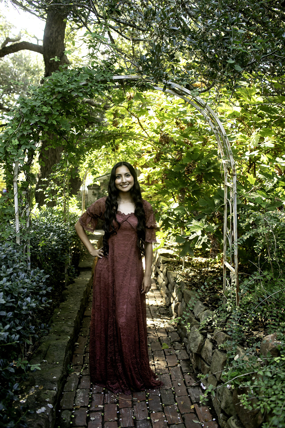 A senior in a flowing gown, standing amidst the lush greenery of Chandor Gardens in Weatherford, TX, radiating confidence and joy.
