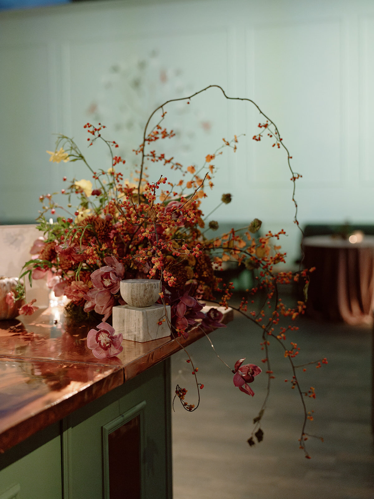 Trailing bar meadows with florals in hues of purple, ruby, copper, gold, and rust for fall wedding reception. Sculptural product clustered floral design for fall wedding. Product clustered floral statements in a tasteful fall color palette of copper, ruby, gold, burgundy, rust, and lavender. Orchids, bittersweet berries, butterfly ranunculus, and tulips are highlighted. Unique floral design with flower forward large scale installations. Design by Rosemary & Finch Floral Design in Nashville, TN.