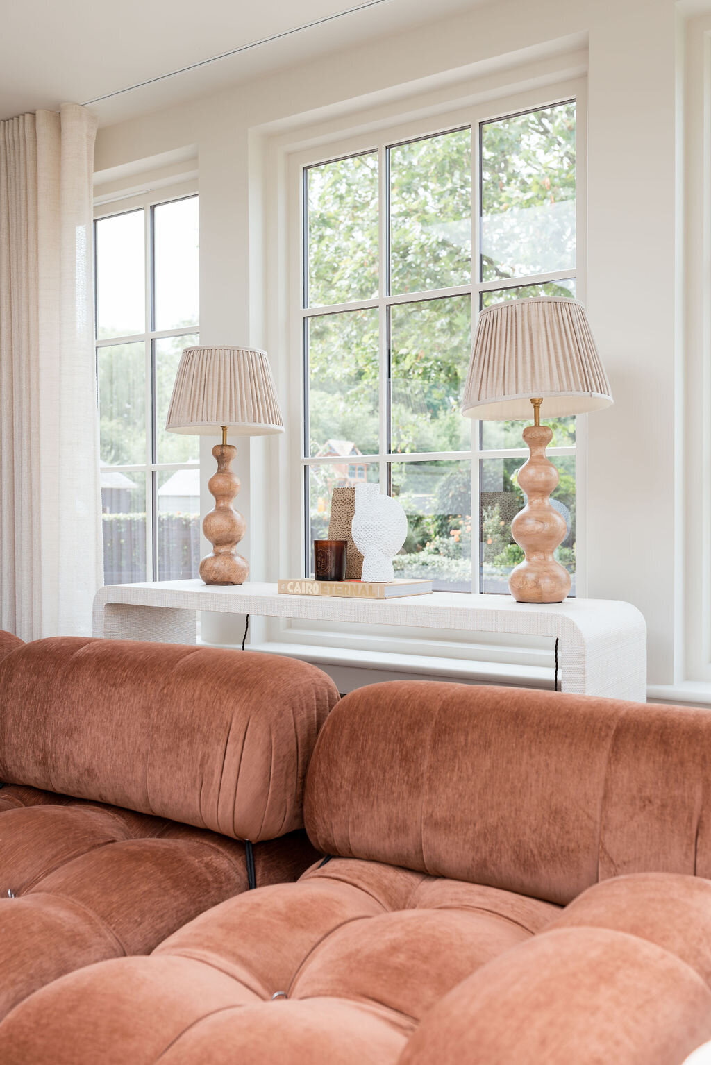 A console table nestled behind a plush, rust orange sofa. Two large neutral lamps sit on the table. Behind the table are lage windows, showing greenery outside.