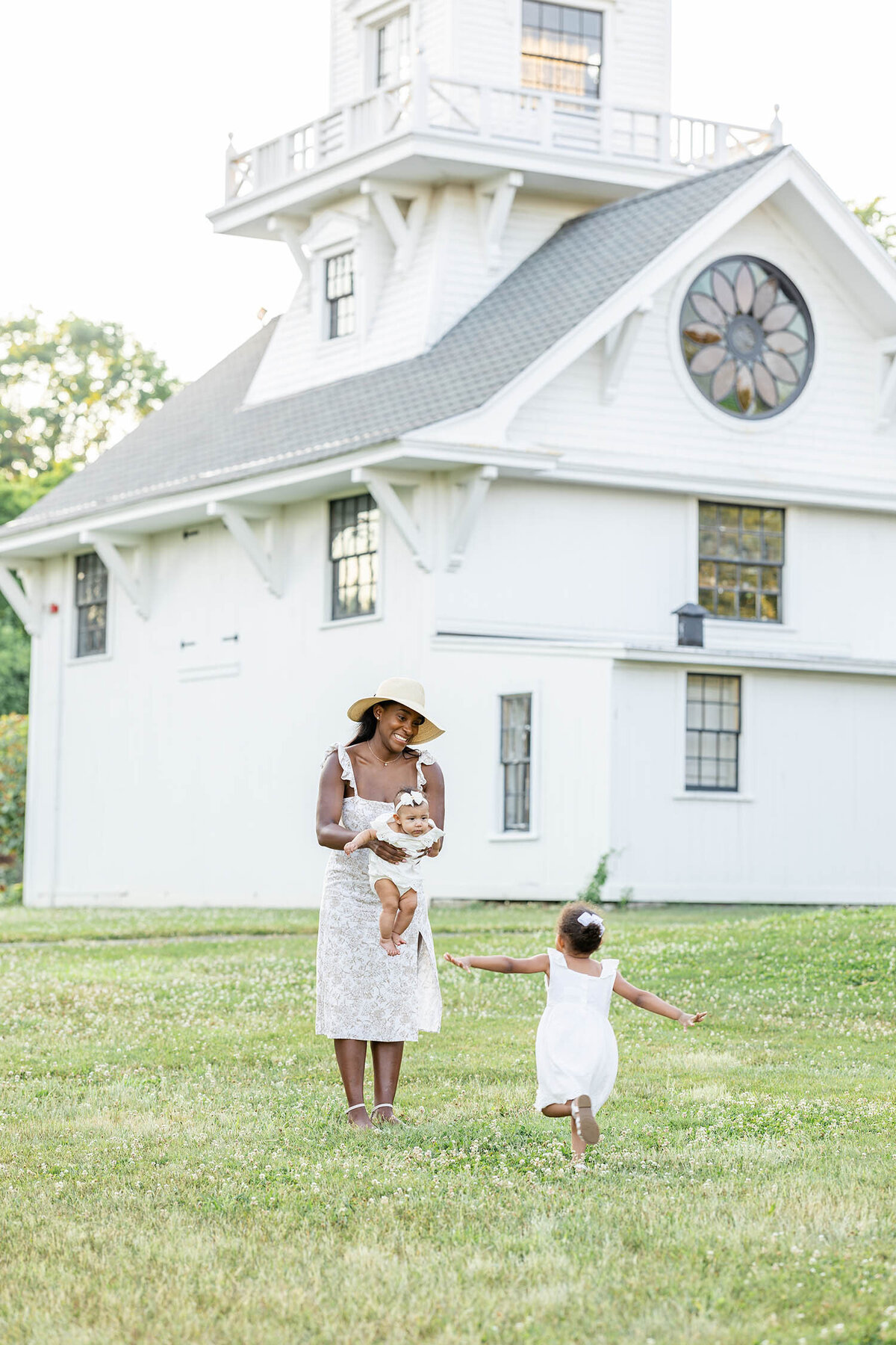Toddler girl runs towards woman who is standing with a baby lowered in her arms in an open clover field during a mommy and me session.