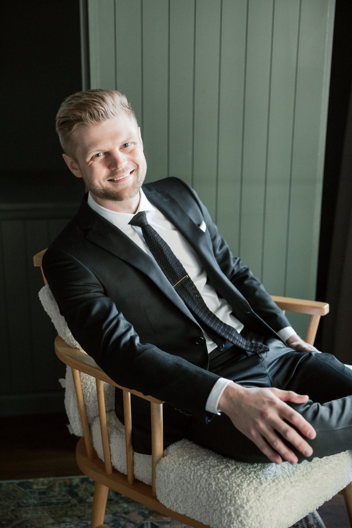 Groom smiling in a black suit, seated indoors with modern decor, taken in Dayton by a Philadelphia wedding photographer.