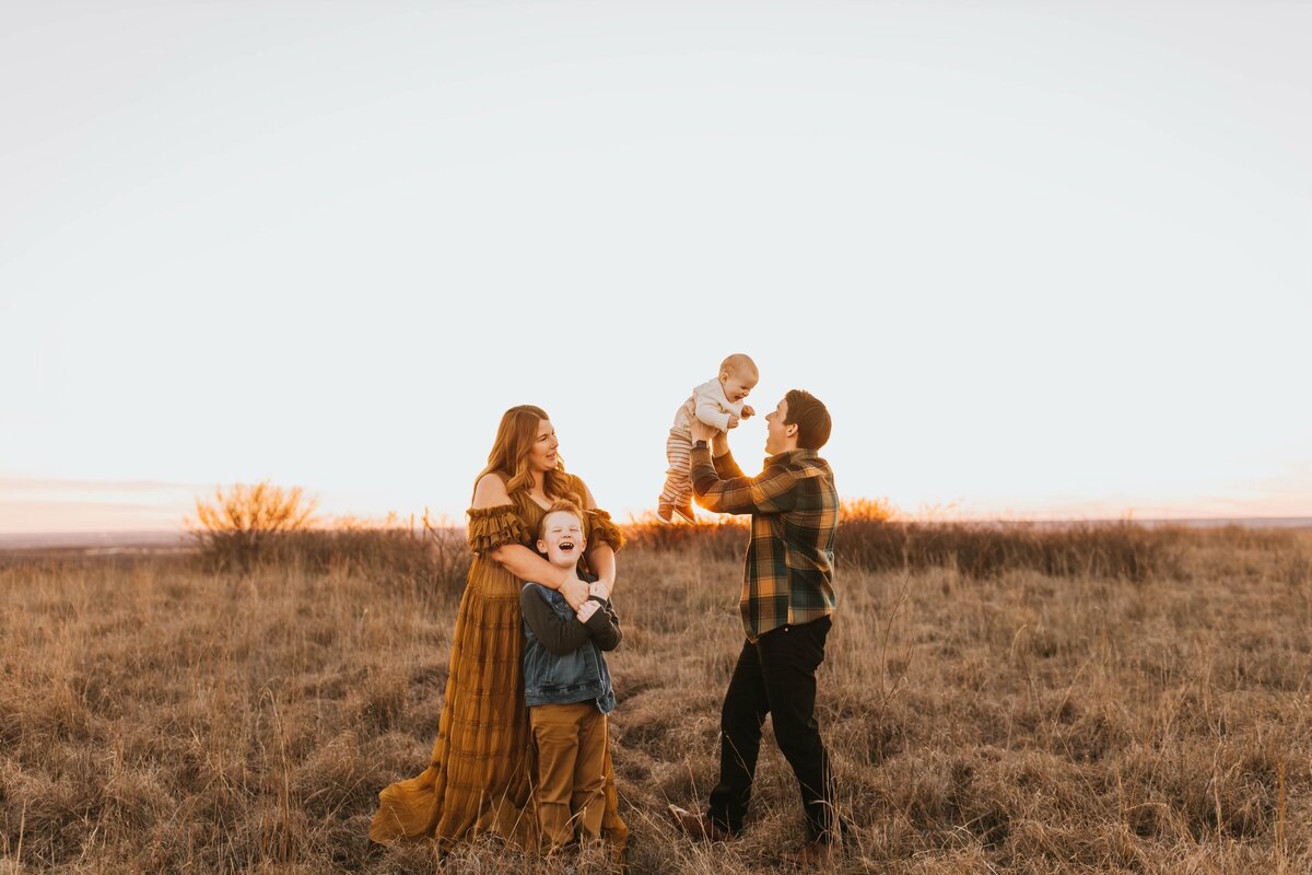 dad hoisting baby in the air while mom and oldest son snuggle up and watch with a smile