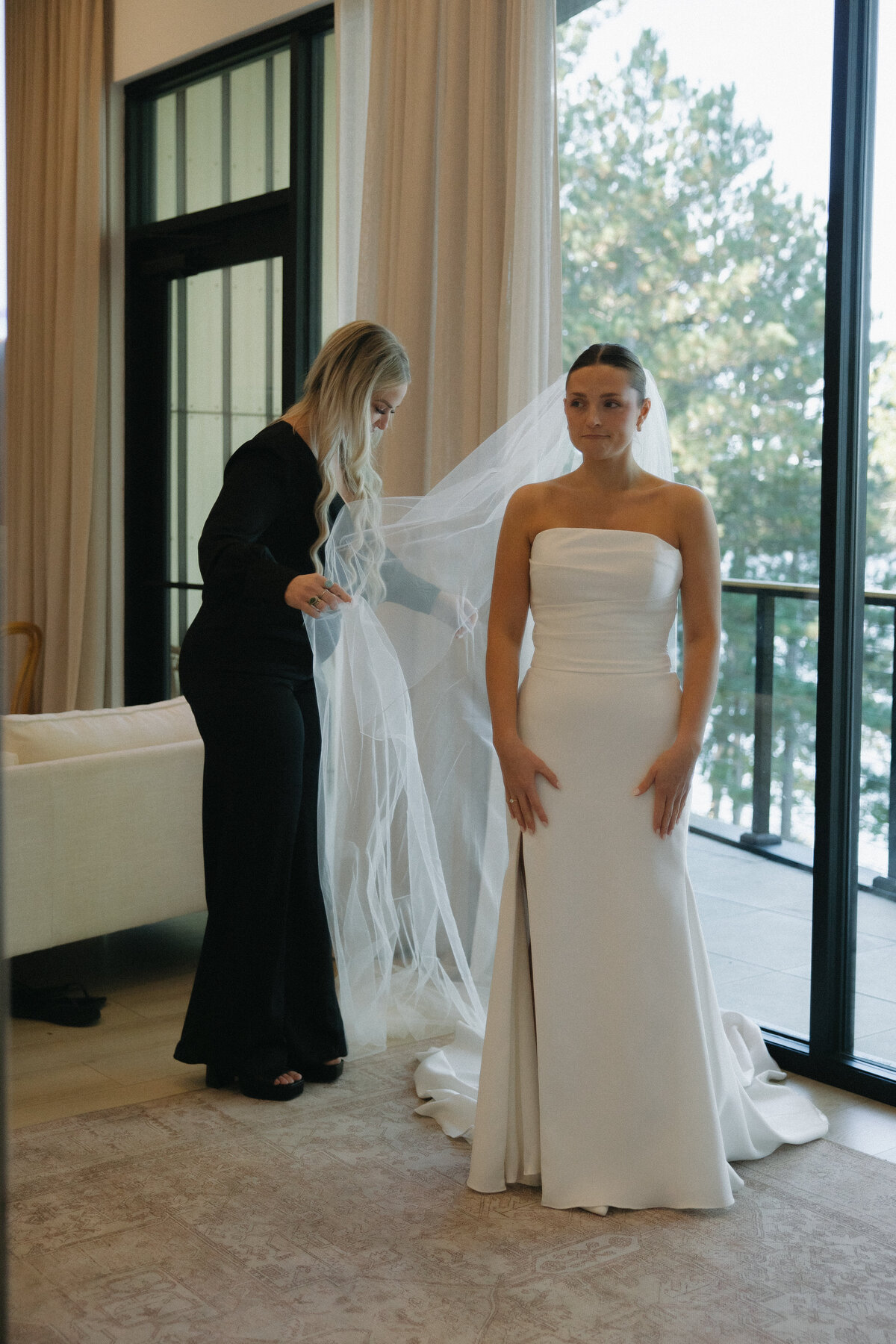 A woman in a black pant suit and platform sandals with long blond hair helps to extend a cathedral length veil out from behind a bride standing in a floor length wedding dress. The dress is white satin and strapless. There are pleats at the top of the dress creating texture to the dress and there is a thigh high leg slit. Behind the duo, there is a large window overlooking trees and a curtain that is the length of the wall. 