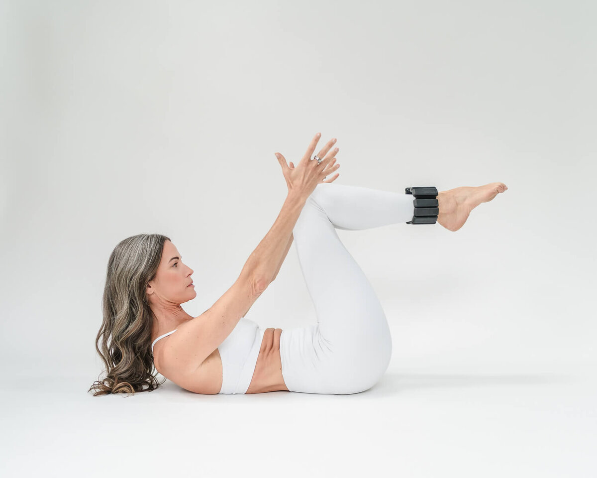 Jenn Pike lying on back in white workout set, arms extended toward legs with ankle weights.