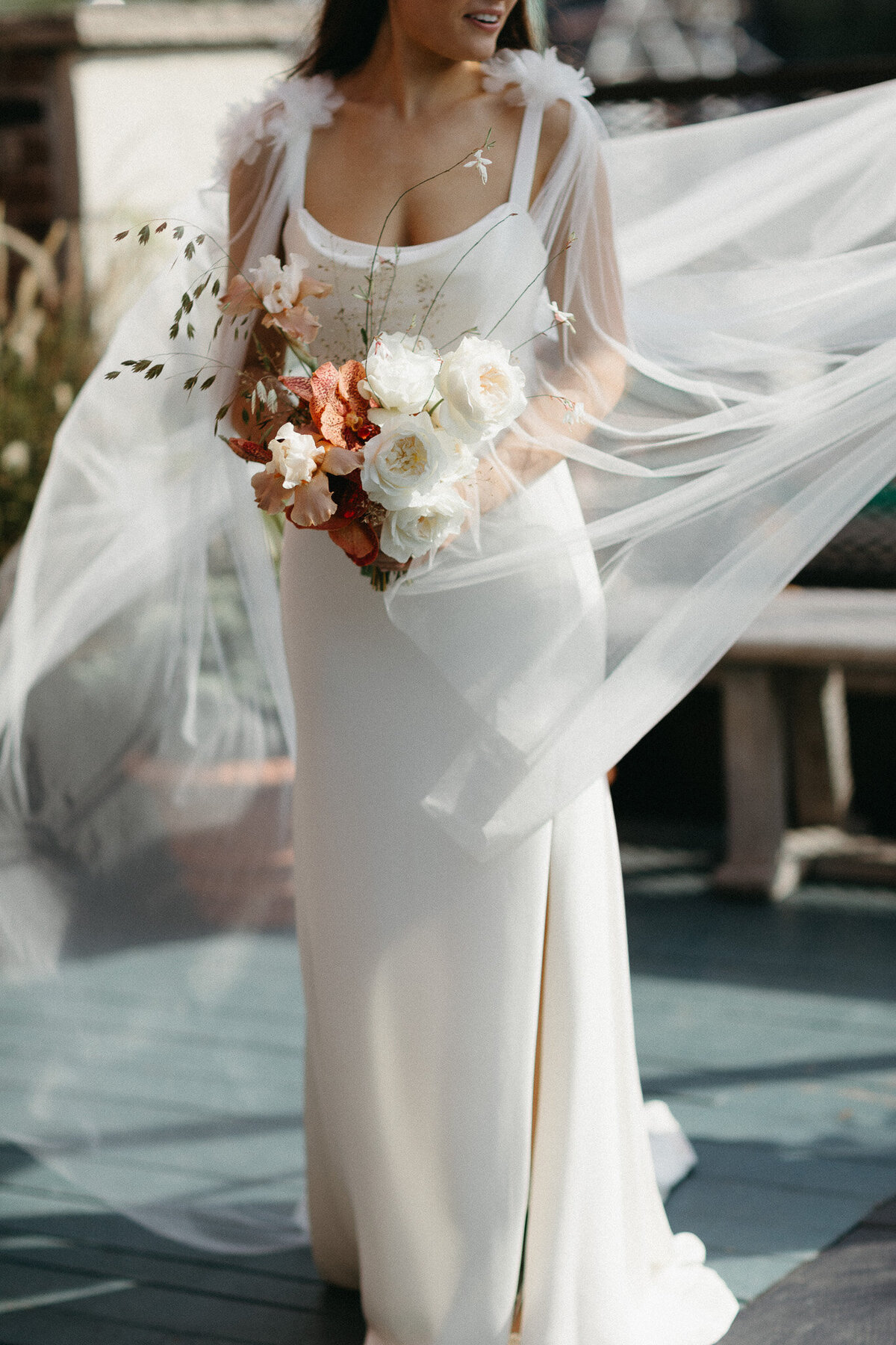 In a graceful white dress with sheer details, the bride holds a bouquet of white and pink flowers as her veil flows in the breeze, beautifully captured by an Oshawa wedding photographer.