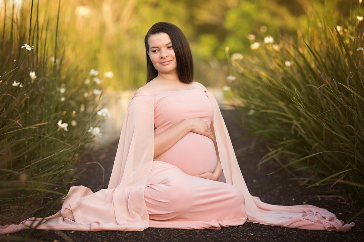 Black woman with straight shoulder length hair sitting in a patch of Louisiana Iris.  She is wearing a pink Silk Fairies Maternity dress.  She is holding her pregnant belly and is in Crescent Park.