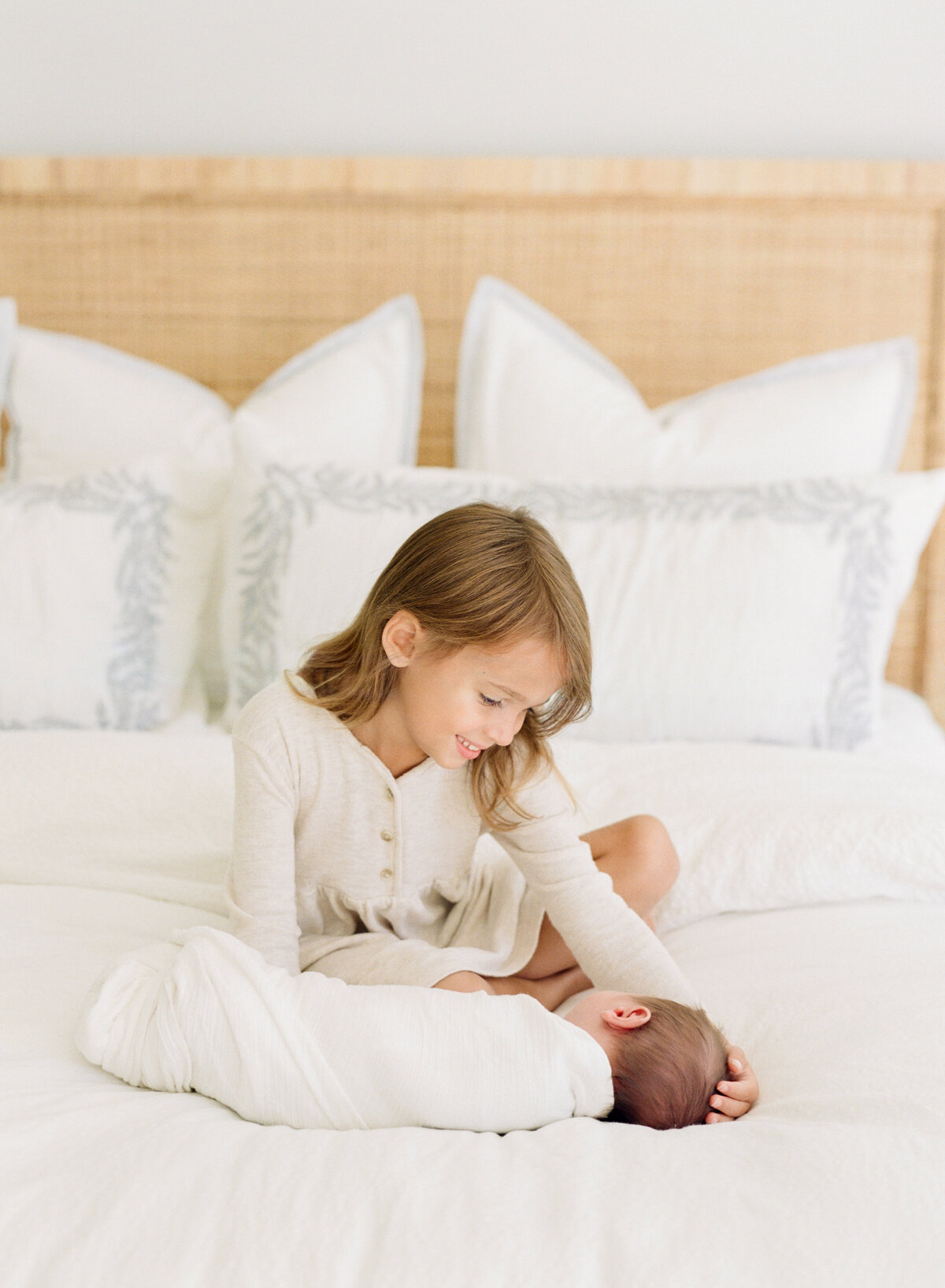 Big sisters sits on a bed with her newborn brother and smiles down at him. Image by Raleigh newborn photographer A.J. Dunlap Photography.