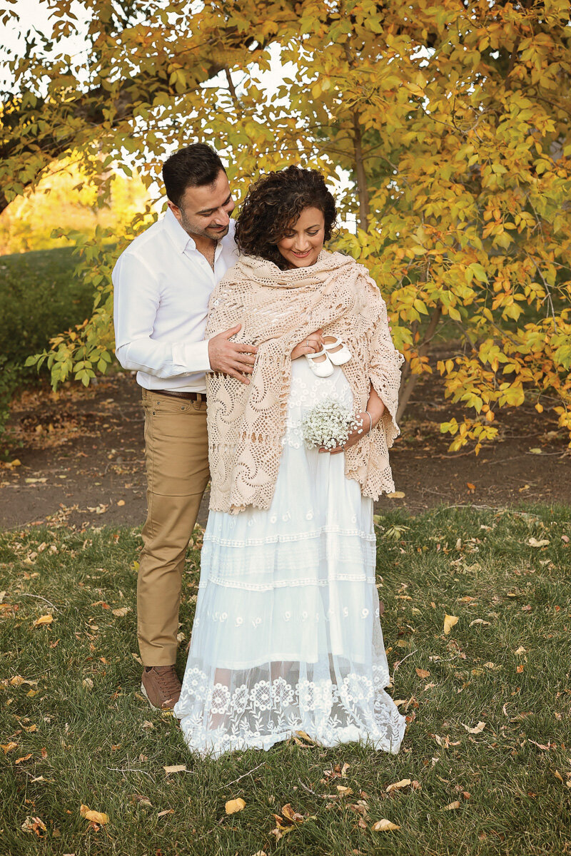 Pregnant woman holding white wild flowers by her pregnant tummy during maternity photo shoot in Calgary Confederation Park by Yuliia Terziian Photography.
